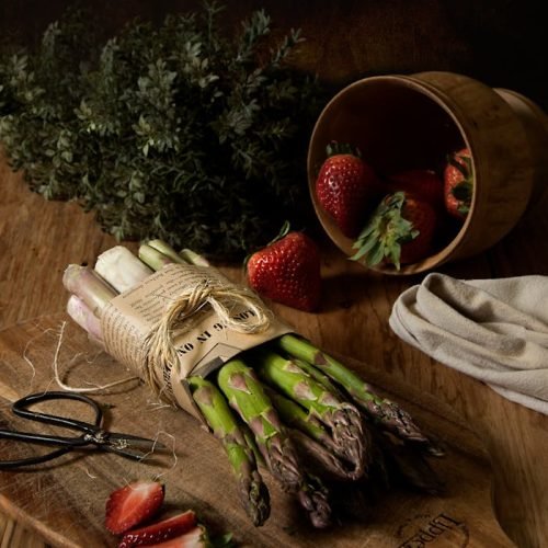 A dark, rustic still life with asparagus and strawberries on a wooden surface.